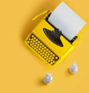 Machine à écrire jaune, sur une surface jaune. Deux boules de papier froissé sont posées à côté.