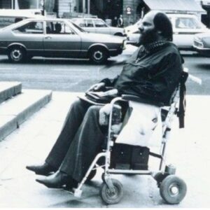 Photos en noir et blanc de Marcel Thorel, un homme chauve et barbu, assis sur un fauteuil électrique rudimentaire chromé, face à un escalier dans la rue.