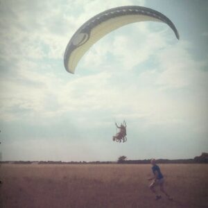 Un parapente en vol sur un ciel cotonneux, peu au dessus du sol. Sont suspendus une personne en fauteuil roulant, et une autre debout derrière elle.